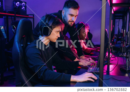 Guy is standing and looking, helping. Gamers are playing video game by computers 136702882