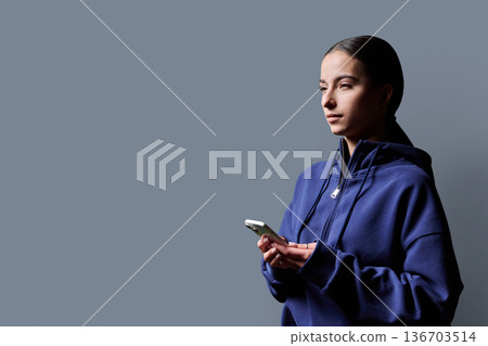 Portrait of serious teenage girl holding smartphone on gray studio background Portrait of serious teenage girl holding smartphone on gray studio background 136703514