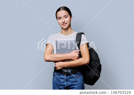 Portrait of beautiful female teenage student with backpack, laptop on gray background 136703529
