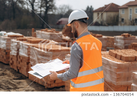 Holding notepad with documents. Construction worker is at job with bricks outdoors Holding notepad with documents. Construction worker is at job with bricks outdoors 136704054