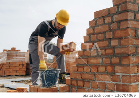 In uniform and hard hat. Construction worker is at job with bricks outdoors In uniform and hard hat. Construction worker is at job with bricks outdoors 136704069
