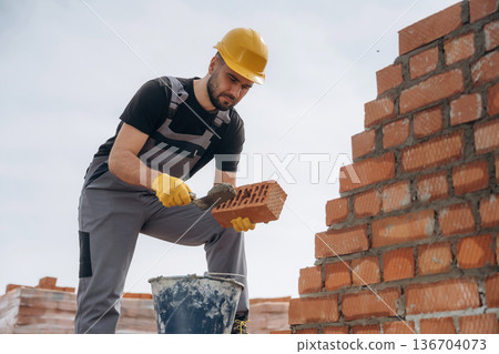 In uniform and hard hat. Construction worker is at job with bricks outdoors In uniform and hard hat. Construction worker is at job with bricks outdoors 136704073