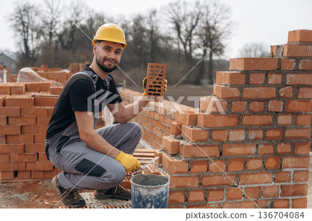 Sitting and building the foundation. Construction worker is at job with bricks outdoors Sitting and building the foundation. Construction worker is at job with bricks outdoors 136704084