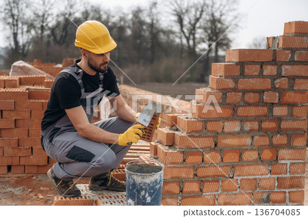 Sitting and building the foundation. Construction worker is at job with bricks outdoors Sitting and building the foundation. Construction worker is at job with bricks outdoors 136704085