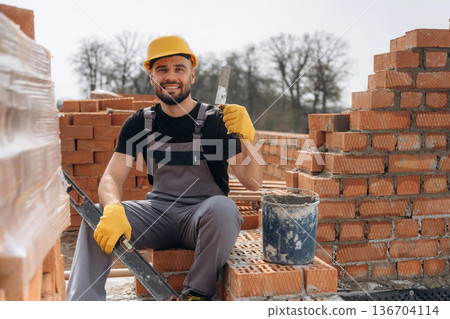 Cheerful positive guy. Construction worker is at job with bricks outdoors Cheerful positive guy. Construction worker is at job with bricks outdoors 136704114