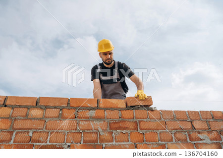 Against cloudy sky, making wall. Construction worker is at job with bricks outdoors Against cloudy sky, making wall. Construction worker is at job with bricks outdoors 136704160