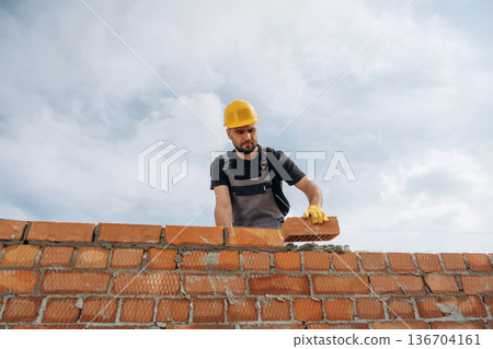Against cloudy sky, making wall. Construction worker is at job with bricks outdoors 136704161