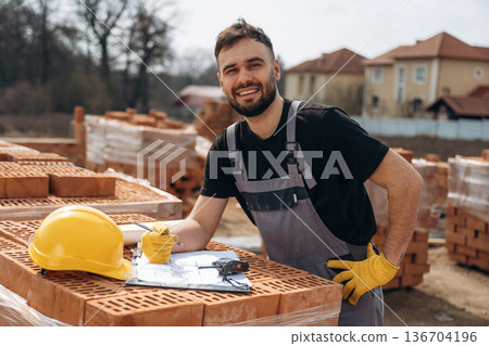 Positive facial expression, standing. Construction worker is at job with bricks outdoors Positive facial expression, standing. Construction worker is at job with bricks outdoors 136704196