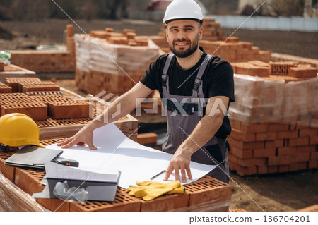 Positive facial expression, standing. Construction worker is at job with bricks outdoors Positive facial expression, standing. Construction worker is at job with bricks outdoors 136704201
