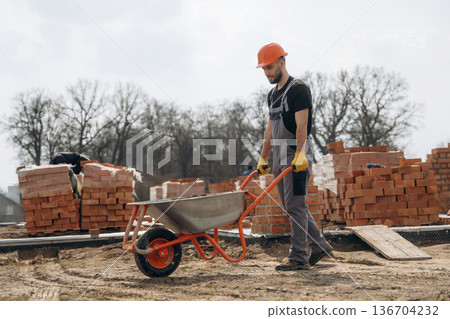 Walking with wheelbarrow. Construction worker is at job with bricks outdoors Walking with wheelbarrow. Construction worker is at job with bricks outdoors 136704232