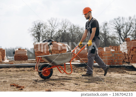 Walking with wheelbarrow. Construction worker is at job with bricks outdoors 136704233