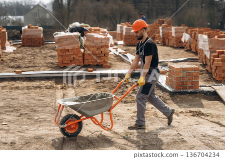 Walking with wheelbarrow. Construction worker is at job with bricks outdoors Walking with wheelbarrow. Construction worker is at job with bricks outdoors 136704234
