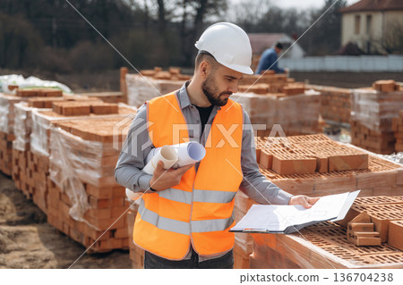 Rolled documents in hand. Construction worker is at job with bricks outdoors Rolled documents in hand. Construction worker is at job with bricks outdoors 136704238