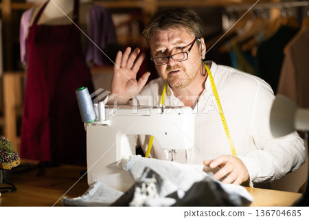 Middle-aged tailor with hearing problems sitting by sewing machine 136704685