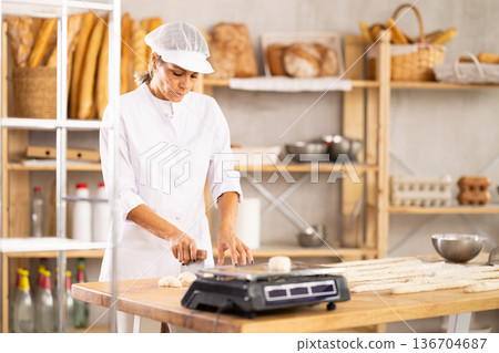 Female baker cutting dough with knife on kitchen table in bakery Female baker cutting dough with knife on kitchen table in bakery 136704687