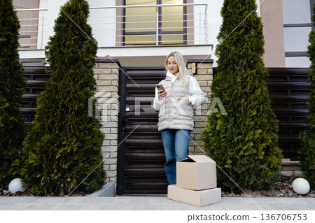 Customer using smartphone waiting for home delivery with cardboard boxes near house 136706753