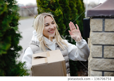 Smiling delivery woman waving while holding a package Smiling delivery woman waving while holding a package 136706758
