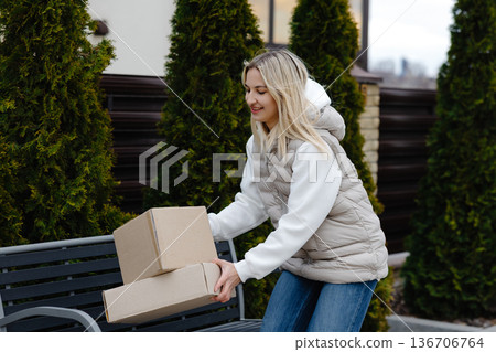 Woman holding cardboard boxes standing next to a bench in front of her house Woman holding cardboard boxes standing next to a bench in front of her house 136706764