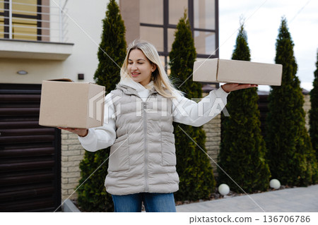 Delivery woman holding two cardboard boxes in front of a house Delivery woman holding two cardboard boxes in front of a house 136706786