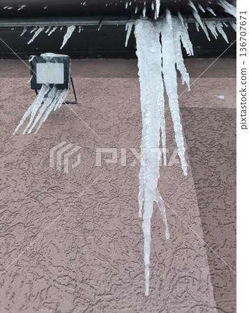Large icicles of ice hang from the gutter of the roof of the house, a spotlight hangs on the wall 136707671