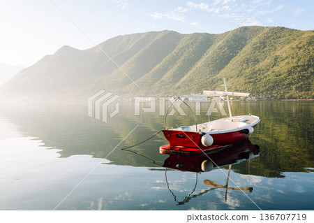 A serene red boat peacefully floating on calm waters surrounded by mountains in early morning light A serene red boat peacefully floating on calm waters surrounded by mountains in early morning light 136707719