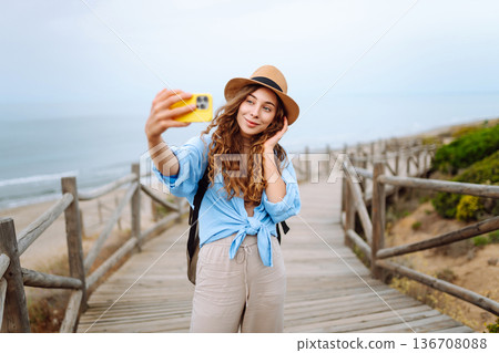 Young woman with curly hair wears a stylish hat and waves while taking a selfie. Travel, tourism 136708088