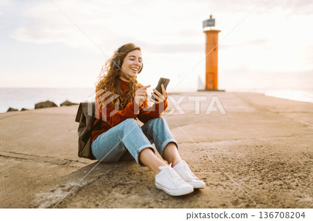 Happy woman listening nice music by waterfront while wearing headphones. Travel, blogging concept. 136708204