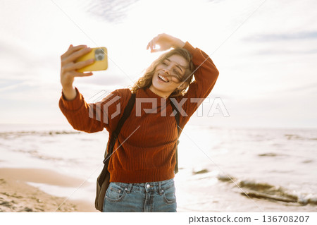 Young woman joyfully taking selfie on beach during golden hour. Selfie time. Cold windy weather. Young woman joyfully taking selfie on beach during golden hour. Selfie time. Cold windy weather. 136708207