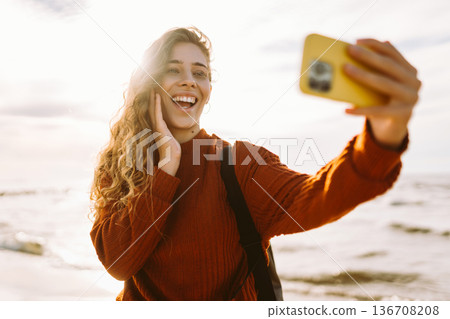 Young woman joyfully taking selfie on beach during golden hour. Selfie time. Cold windy weather. Young woman joyfully taking selfie on beach during golden hour. Selfie time. Cold windy weather. 136708208