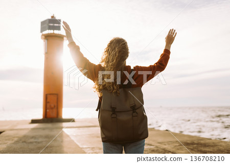 Woman enjoying breezy day near vibrant red lighthouse at sunset. Travel, weekend, lifestyle concept. 136708210