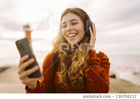 Happy woman listening nice music by waterfront while wearing headphones. Travel, blogging concept. Happy woman listening nice music by waterfront while wearing headphones. Travel, blogging concept. 136708212