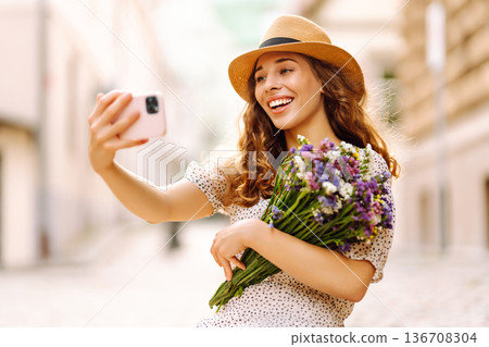 Selfie time. Young woman with a bouquet of wild flowers posing on the street. Fashion style. 136708304