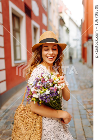 Woman with curly hair smiles while holding bouquet of flowers in city on sunny day. Fashion style Woman with curly hair smiles while holding bouquet of flowers in city on sunny day. Fashion style 136708309