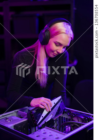 Female gamer builds gaming PC under purple lights with headphones on and focused expression Female gamer builds gaming PC under purple lights with headphones on and focused expression 136708554