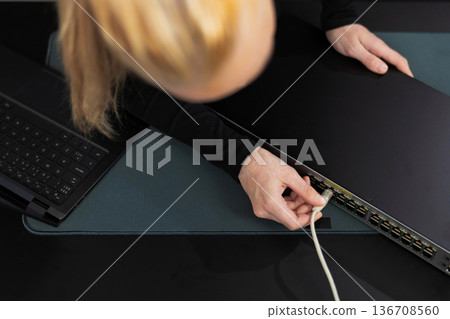 Woman in her 30s with blond hair installs Ethernet cable to network switch in a modern office setting Woman in her 30s with blond hair installs Ethernet cable to network switch in a modern office setting 136708560