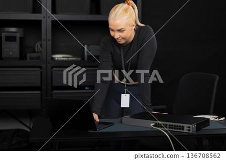 Female IT technician, young adult, blonde, working with a laptop and network switch in a tech office 136708562