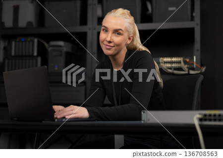 Confident female IT technician at a desk with a laptop and network gear in a technical office 136708563