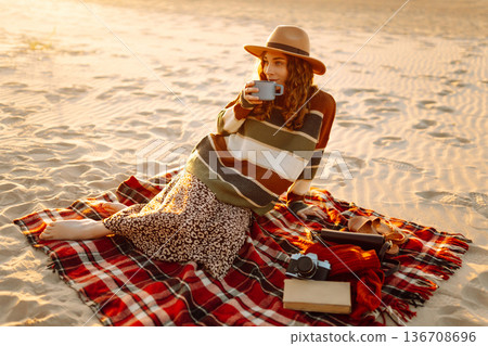 Cute woman sits on the beach drinks a hot drink from a thermos. Picnic, weekend, relax and lifestyle 136708696
