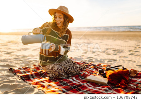 Young woman sits at picnic on the beach drinks a hot drink from a thermos. Relax and lifestyle 136708742