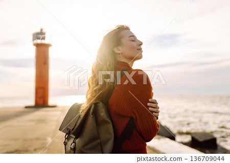 Young woman enjoying a breezy day near a vibrant red lighthouse. Autumn sunset Young woman enjoying a breezy day near a vibrant red lighthouse. Autumn sunset 136709044