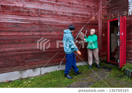 After hanging Christmas garland on house facade man carries ladder inside. 136709165