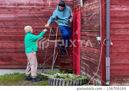 During Christmas eve garland installation on wall of rural house woman passes nails to man. 136709169