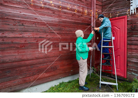 Man and woman over 50 years old are hanging garland on external wall of rural house. 136709172