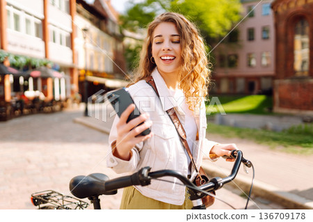 Woman using mobile phone, walks with bicycle, looks around at beautiful architecture. Selfie time. 136709178