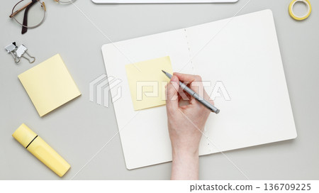 Woman writing on sticky note at organized workspace with notebook and stationery items 136709225
