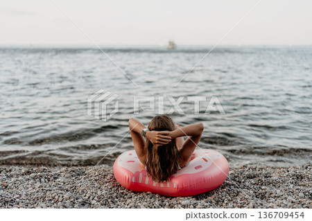 Woman, beach, relaxing. Young woman relaxing on a donut float at a pebble beach enjoying summer vacation. Woman, beach, relaxing. Young woman relaxing on a donut float at a pebble beach enjoying summer vacation. 136709454