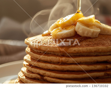Steaming stack of wholegrain pancakes topped with fresh banana slices and pouring honey in warm morning sunlight. 136710102