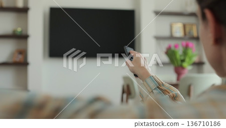 Woman selects tv channels with remote control at living room on sofa. Woman controls TV using a modern remote control. 136710186