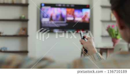 Woman selects tv channels with remote control at living room on sofa. Woman controls TV using a modern remote control. Woman selects tv channels with remote control at living room on sofa. Woman controls TV using a modern remote control. 136710189
