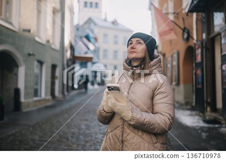 Woman looks up while holding phone in an old city street during winter morning Woman looks up while holding phone in an old city street during winter morning 136710978